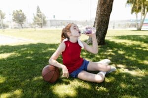 Girl in red tank top and blue shorts drinking water on grass. Learn about water dehydration and survival without water or food.