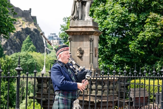 A Scottish bagpiper dressed in a traditional kilt and blue jacket playing the Great Highland Bagpipes near a historic monument in Edinburgh