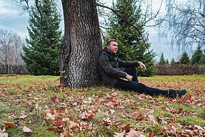 Man in a jacket sitting under a tree in an autumn park, surrounded by leaves