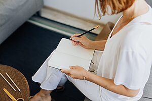 A person sitting and writing in a travel journal on a cozy indoor sofa setting.