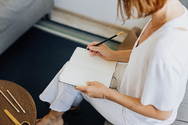 A person sitting and writing in a travel journal on a cozy indoor sofa setting.