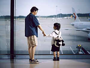 Single parent and child holding hands, gazing at planes through an airport window.