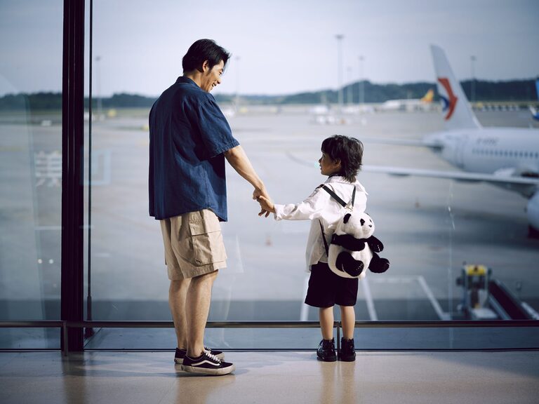 Single parent and child holding hands, gazing at planes through an airport window.