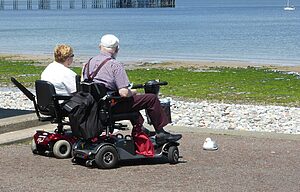 Elderly couple enjoys a seaside view while seated on mobility scooters, emphasising leisure and accessibility.