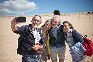 A joyful group of senior friends taking a selfie at a sandy beach in Portugal under clear blue skies.