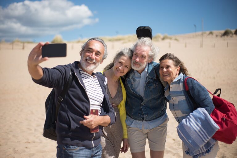 A joyful group of senior friends taking a selfie at a sandy beach in Portugal under clear blue skies.