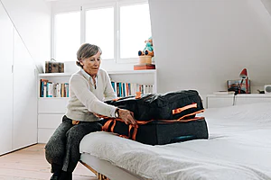 Elderly woman packing suitcase on bed in bright, modern bedroom setting.