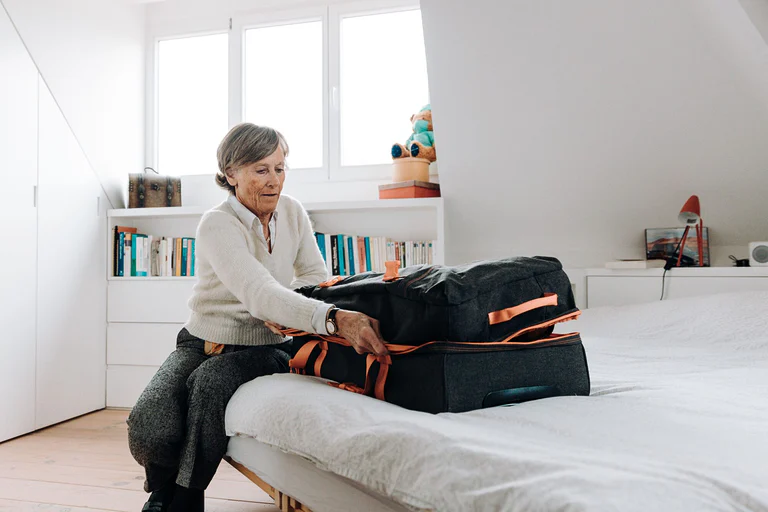 Elderly woman packing suitcase on bed in bright, modern bedroom setting.