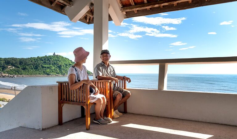 Senior couple sitting on balcony chairs, enjoying a relaxing ocean view in summer.