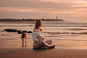 Pregnant woman sitting on a beach next to a dog.