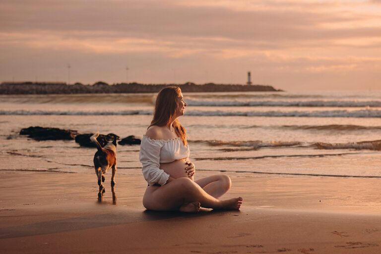 Pregnant woman sitting on a beach next to a dog.