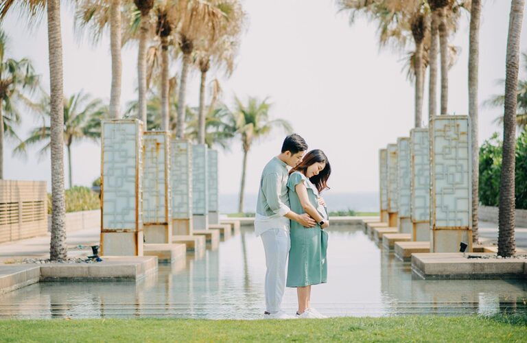 Pregnant couple embraces amidst palm trees reflecting tranquillity and love.