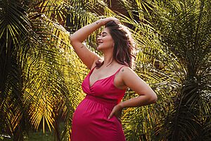 A pregnant woman in a vibrant pink dress poses happily among lush green plants outdoors.