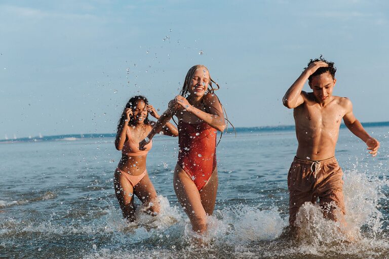 Three teenagers enjoy a playful day at the seaside, splashing through the waves under a clear summer sky.